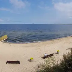 Spokojna plaża dla psów z piaskiem i morzem. Na ławce siedzi osoba, obok kosze.