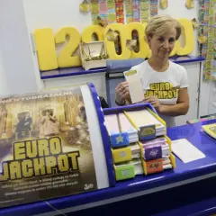 Kobieta w koszulce Eurojackpot trzyma kupon na tle stoiska z grami losowymi.