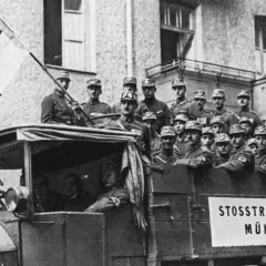 Männer in Uniformen auf einem LKW mit der Aufschrift "STOSSTRUPP HITLER MÜNCHEN". Ein Mann hält eine Fahne mit einem Adler. Dies ist ein Bild vom Hitlerputsch.