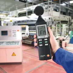 Mano sostiene sonómetro que mide 81.7 dB(A) en una fábrica, indicando el nivel de ruido.