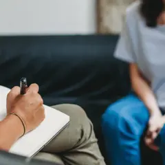 Eine Person schreibt in ein Notizbuch, während eine andere Person in blauen Jeans auf einer Couch sitzt. Dies könnte eine Szene der MDMA-Therapie sein, die bei der Heilung hilft.