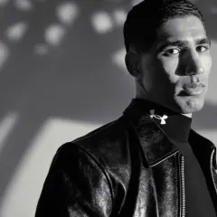 Achraf Hakimi, ein Fußballstar, blickt nachdenklich in die Kamera, trägt eine Lederjacke und ein schwarzes Rollkragenshirt mit dem Under Armour Logo.