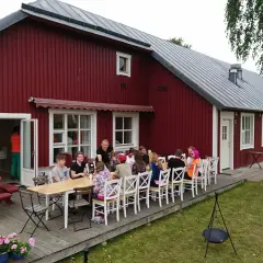 Gästerna njuter av en måltid utomhus vid Sjötorp vandrarhem. En grupp vänner samlas runt borden på altanen framför den röda byggnaden.