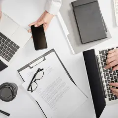 Dwie osoby pracują przy biurku, używając laptopów i telefonu. Na stole leżą dokumenty, okulary, lupa i kubek. Sprawdzają, ile widnieje się w BIK.