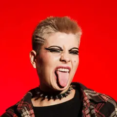 Młoda osoba z blond włosami i mocnym makijażem oczu, w kolczatce i flanelowej koszuli, wyciąga język. To kwintesencja buntu i stylu punk.
