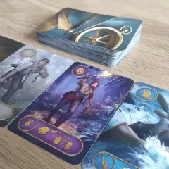 Na stole leżą karty z gry ryzykanci. Widoczny jest stos kart z kompasem, a obok cztery karty przedstawiające postacie: starszego pana z fajką, mężczyznę z aparatem, kobietę z pochodnią i mężczyznę w łodzi na rwącej rzece.
