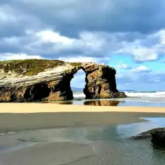 Arco natural en las playas de Galicia, con arena dorada, mar azul y cielo nublado.