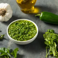 Miseczka pełna aromatycznego sosu chimichurri, obok świeża kolendra, natka pietruszki, czosnek, papryczka jalapeno i oliwa.