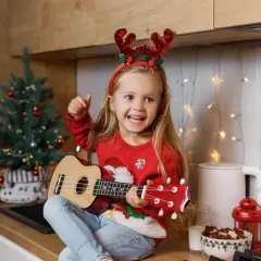 Dziewczynka w świątecznych rogach renifera gra na ukulele w kuchni, obok choinki i lampek.