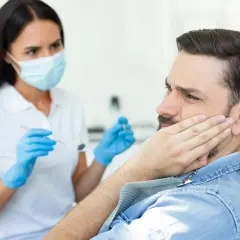 Mężczyzna z bólem policzka u dentysty, który bada jego dziąsła. Może to być objaw pogłębiających się kieszonki dziąsłowe.