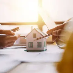 Mani che studiano un modello di casa, con matite e calcolatrice. Si discute il surroga mutuo significato per un nuovo acquisto immobiliare.