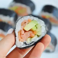 Ręka trzyma kawałek futomaka z łososiem i awokado. Na talerzu widać więcej sushi.