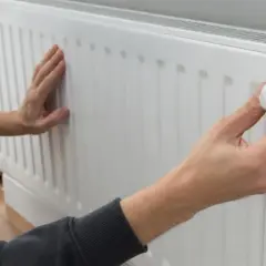Ręce dotykają białego grzejnika, sprawdzając, czy jest zimny. Jedna ręka reguluje termostat.