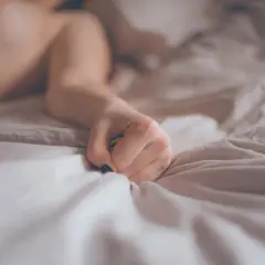 A person's hand clutches crumpled white sheets on a bed.