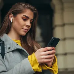 Kobieta z białymi słuchawkami bezprzewodowymi w uchu sprawdza coś na telefonie. Dowiedz się, jak włączyć słuchawki bezprzewodowe.