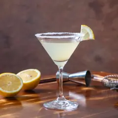 Elegancki martini bianco drink z solnym rantem i plasterkiem cytryny, obok połówki cytryny, oliwki i akcesoria barmańskie.