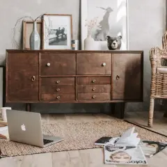 Stary, drewniany kredens z tekowe drewno, obok wiklinowy fotel. Na dywanie laptop, książki i czasopisma.