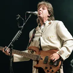 Paul McCartney z zespołu The Beatles gra na basie podczas koncertu, ubrany w elegancki beżowy garnitur.
