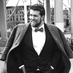 Un homme gros bien habillé, souriant, porte un costume sombre, une chemise blanche et un nœud papillon. Son manteau est jeté sur ses épaules.