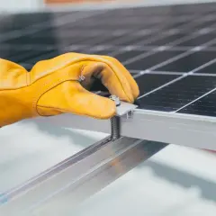 Ręka w żółtej rękawicy dokręca śrubę mocującą panel fotowoltaiczny. Instalacja PV postępuje.