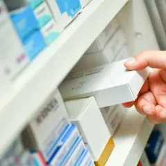 Dłoń sięga po białe pudełko leku, być może zawierającego nilogrin, z aptecznej półki pełnej innych opakowań.