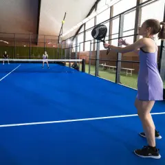 Ragazza in abito viola gioca a padel, con racchetta in mano, su campo blu. Altri giocatori in lontananza.