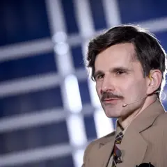 Mężczyzna w garniturze i z wąsami, z mikrofonem przy uchu, na tle niebiesko-białych pasów.