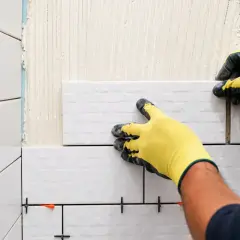 Pose de faience murale. Mains gantées en jaune plaçant une nouvelle tuile sur un mur préparé avec du mortier.