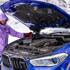 Mężczyzna w fioletowej kurtce podłącza booster do akumulatora niebieskiego BMW. To rozwiązanie, co może pobierać prąd w samochodzie na postoju, gdy jest zimno.