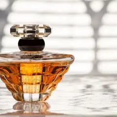 Elegancka butelka perfum o bursztynowym kolorze, z kryształowym korkiem, stoi na błyszczącej powierzchni.