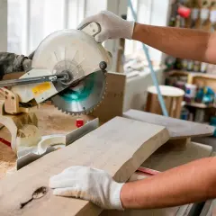 Un artisan utilise une scie à onglet pour couper une planche de bois. Il tient la pièce avec des gants, prêt à réaliser une coupe précise.