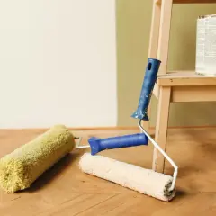Färgburk och färgroller redo att måla på tapet. En stege står i bakgrunden.