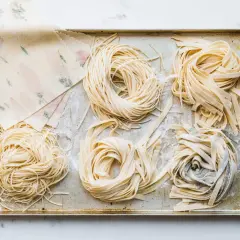 Frische Nudeln in verschiedenen Formen liegen auf einem Blech. Ein Stapel mit Blumenmuster verzierter Teigblätter liegt daneben. Perfekt für alle, die Nudeln selber machen wollen.
