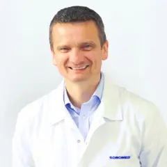 Uśmiechnięty mężczyzna w białym fartuchu lekarskim, z niebieską koszulą pod spodem.