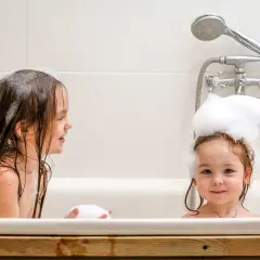 Dwie dziewczynki w wannie, jedna z pianą na głowie, druga z mokrymi włosami. Radosne mycie głowy dziecku to świetna zabawa.