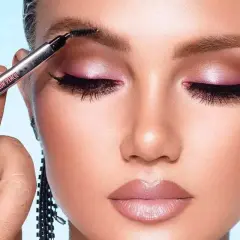 Occhi con ombretto rosa scintillante e labbra morbide. Una mano sfuma le sopracciglia con un pennellino, completando un look da sogno.