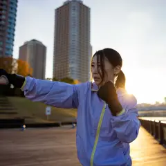 A woman practices shadow boxing outdoors, a popular fitness trend, with city buildings in the background at sunset.