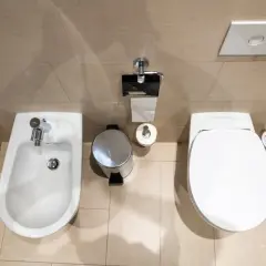 Łazienka z bidetem i WC. Widać kosz na śmieci, papier toaletowy i szczotkę. Odległość WC od ściany bocznej jest optymalna.