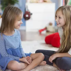 Dwie dziewczynki siedzą na dywanie, rozmawiając. To świetny sposób, jak zachęcić dziecko do mówienia w przedszkolu.