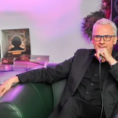 Marek Biliński, w czarnym garniturze, siedzi na zielonej kanapie, trzymając mikrofon. W tle widać płyty z jego utworami.