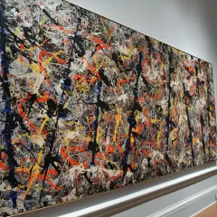 A woman admires a massive Jackson Pollock painting worth $140 million, a chaotic symphony of splattered paint in black, white, orange, and blue.