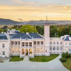 Elegancki zamek na Węgrzech, otoczony zielenią, z wieżą i łukowymi oknami, na tle zachodzącego słońca.