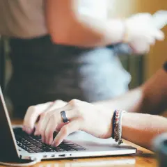 Dłonie piszą na laptopie, przygotowując księgowanie alior. W tle widać inne osoby i roślinę.