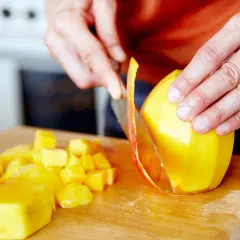 Hände schneiden eine reife Mango auf einem Holzbrett. Saftige Fruchtstücke liegen daneben.