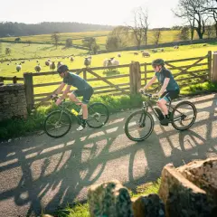 Two cyclists enjoy a scenic ride, cycling in the Cotswolds with sheep grazing in a sun-drenched field beside a stone wall.