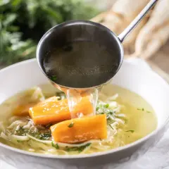Un mestolo versa brodo caldo in una ciotola di zuppa di pollo con carote e tagliatelle.