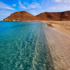 Aguas cristalinas acarician la arena dorada de las playas vírgenes de Almería, con montañas áridas de fondo bajo un cielo azul.