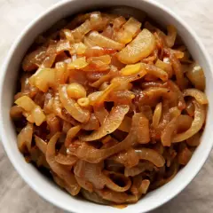 Un bol rempli d'oignons caramélisés dorés, parfaits pour une recette savoureuse. Un oignon cru à côté.