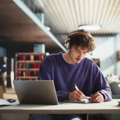 Ein junger Mann mit Kopfhörern lernt in einer Bibliothek. Er schreibt Notizen, während sein Laptop geöffnet ist. Perfekt für ein Teilzeitstudium.