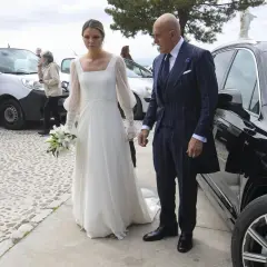 Novia con ramo de lirios y su acompañante, un hombre con traje de raya diplomática, llegan a su boda. Irene Matamoros, radiante.